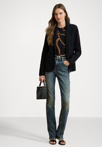 Zwarte blazer over een zwart T-shirt met grafische print en gouden opdruk, gecombineerd met vervaagde blauwe jeans, een zwarte handtas en zwarte sandaaltjes met open tenen.