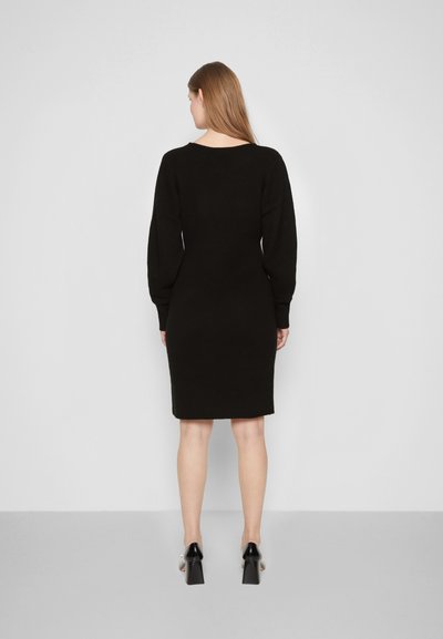 Vestido de malha preto com mangas longas, silhueta ajustada e decote redondo. Textura suave com bainha de comprimento médio, usado com sapatos de salto bloco.