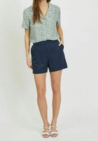 Chemise à manches courtes avec col, motif floral bleu, vert et jaune, associée à un short bleu marine et des sandales blanches.