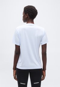 Άτομο με κοντά, σγουρά μαλλιά που φοράει ανοιχτό μπλε κοντομάνικη μπλούζα και μαύρο παντελόνι με ανακλαστικές ρίγες, στάσιμο με την πλάτη προς τα εμάς.