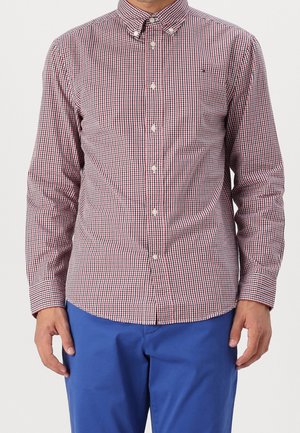 Man wearing long-sleeve red, white, and blue small checkered button-up shirt paired with bright blue pants, standing against plain background.