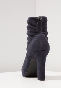 Bottine en daim bleu marine avec tige souple, languette de tirage à l'arrière et talon haut épais, posée sur une surface blanche.