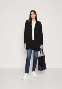 Sort åben front cardigan, hvid skjorte, mørkeblå jeans, hvide sneakers og en sort tote bag. Blød tekstur med afslappet pasform.