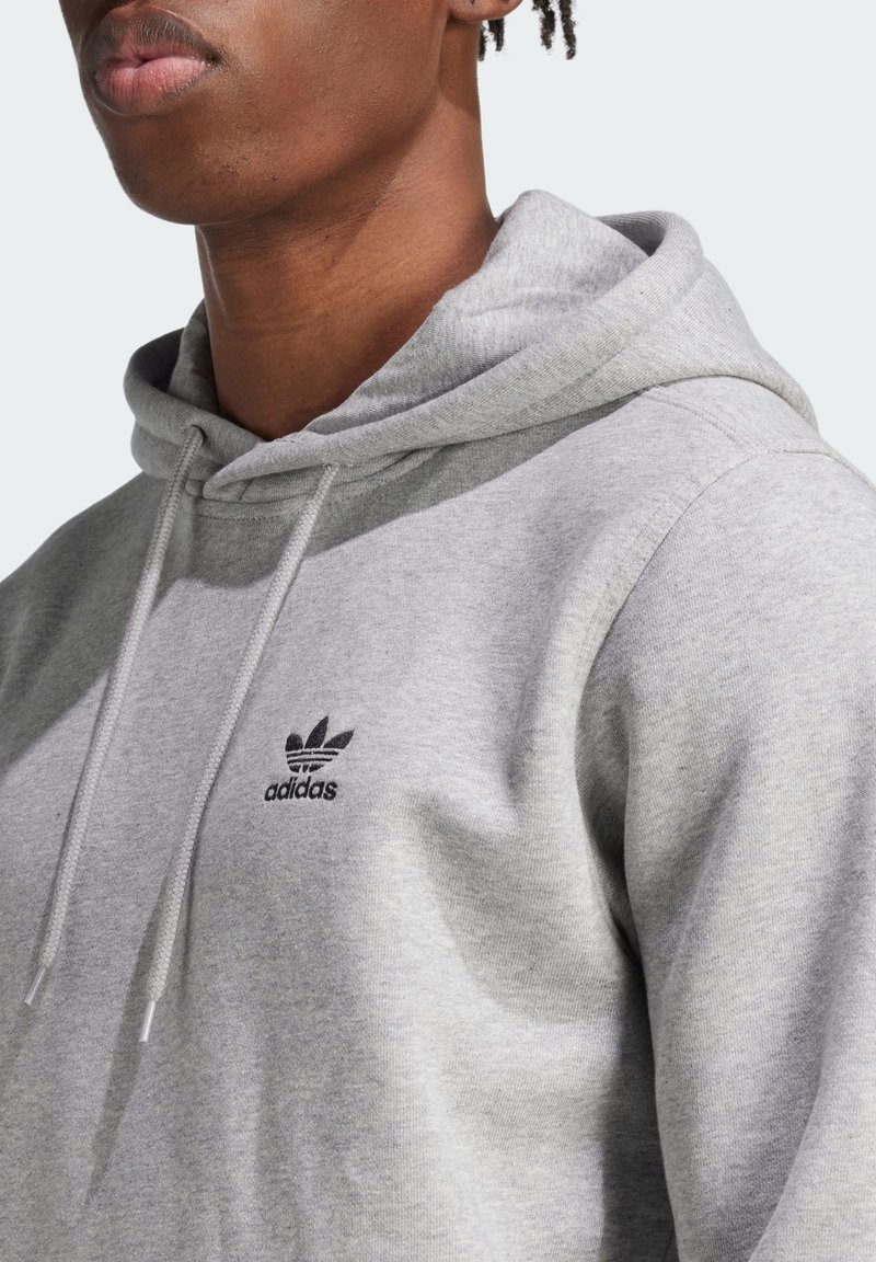 Szara bluza z kapturem wykonana z miękkiego materiału, z czarnym haftowanym logo Adidas na klatce piersiowej i sznurkami ze metalowymi końcówkami.