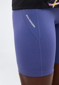 Κοντινή λήψη μωβ αθλητικών σορτς με ορατές ραφές και λευκό λογότυπο "Salomon" στο πλάι του μηρού.