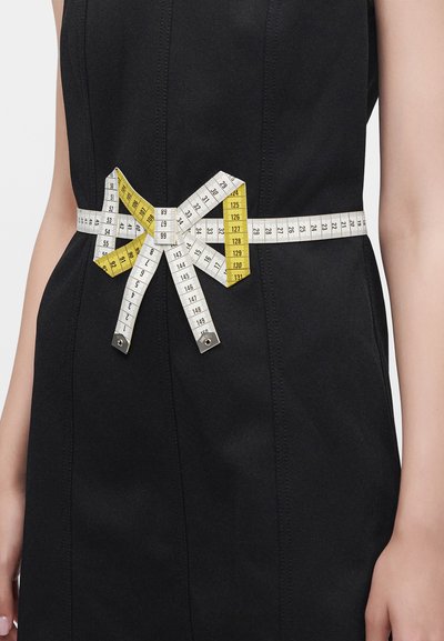 Mètre ruban noué en nœud autour de la taille d'une personne portant une robe noire sans manches, mettant en valeur la zone de la taille.