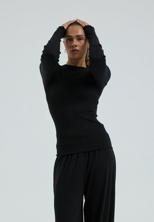 Femme aux cheveux bruns, vêtue d'un haut à manches longues noir et d'un pantalon noir, debout avec les mains posées sur la tête, devant un fond gris uni.