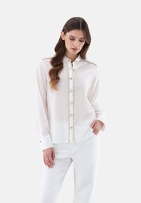 White long-sleeve shirt with a button-down front, gold hardware accents, and a textured collar detail. Worn with white pants.
