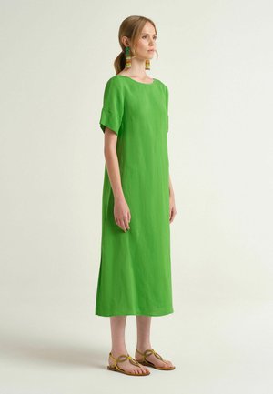 Donna in piedi, che indossa un vestito midi verde brillante con maniche corte, orecchini colorati con perline e sandali gialli bassi.