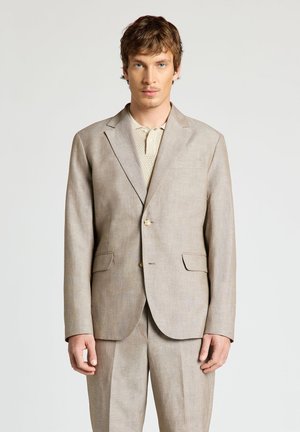 Homme portant un costume en lin beige clair avec un blazer à deux boutons et un pantalon assorti, debout devant un fond clair uni.