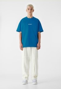 Blå bomulds T-shirt med hvid tekst "LOST YOUTH", parret med cremede teksturerede bukser. Modellen står i hvide sneakers mod en hvid baggrund.