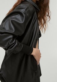 Camicia giacca oversized in pelle nera con texture liscia, chiusura a bottoni e polsini a coste. Presenta una vestibilità rilassata e un colletto ampio.