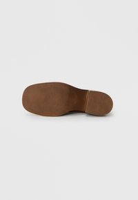 Semelle de chaussure en cuir marron avec un grip texturé, une forme arrondie lisse et un détail de branding minimal embossé à la surface.