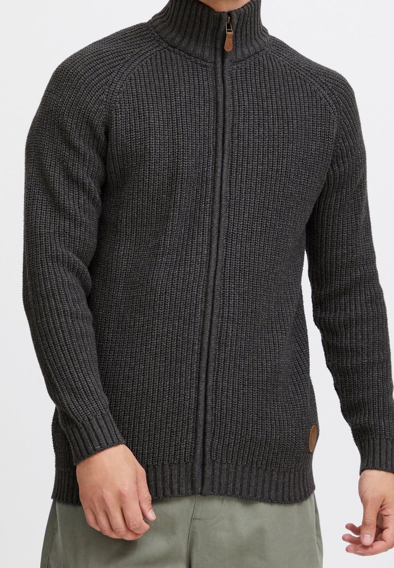 Pull en maille gris foncé à fermeture éclair intégrale avec col montant porté par une personne, associé à un pantalon gris clair.