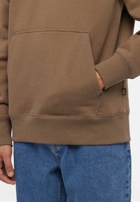 Felpa a girocollo marrone con tasca frontale, polsini e orlo ribattuti, realizzata in tessuto morbido, abbinata a jeans di denim blu.