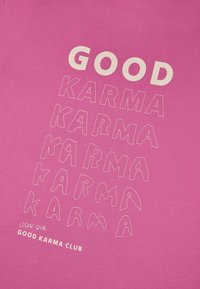 Camiseta rosa con letras audaces y estilizadas. "GOOD" en color blanco sucio en la parte superior, seguido de "KARMA" en capas con diferentes estilos y tamaños.