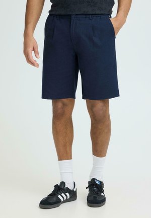 Uomo con pantaloncini blu navy, calzini bianchi e scarpe da ginnastica Adidas nere con strisce bianche, in piedi con una mano in tasca.