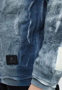 Pull tricoté bleu clair avec motifs torsadés détaillés, poignets côtelés et deux étiquettes de marque visibles sur le côté et la manche.