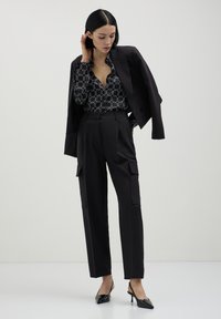 Blazer nero su misura sopra una blusa nera a fantasia, abbinato a pantaloni cargo neri a vita alta e décolleté neri con tacco. Tessuto morbido.