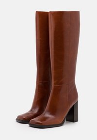 Bottes en cuir marron jusqu'aux genoux avec un bout carré, finition brillante et un large talon noir. Présente des détails de couture verticaux et une fermeture éclair sur le côté.