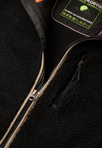 Giacca in fleece nera con zip frontale, caratterizzata da rifiniture beige a contrasto, dettagli della zip visibili e un'etichetta verde all'interno.