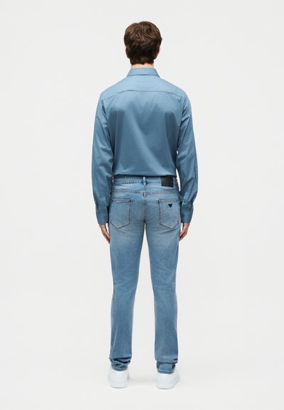 Άνδρας που στέκεται με την πλάτη προς τα εμπρός, φορώντας γαλάζιο πουκάμισο με μακριά μανίκια, γαλάζια τζιν με πίσω τσέπες και λευκά αθλητικά παπούτσια με φόντο απλό.