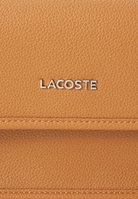 Tan-färgad läderplånbok med sydda detaljer och en metallisk "LACOSTE"-logotyp. Kompakt design med en slät yta och en tydlig form.