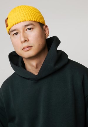 Młody mężczyzna w jaskrawożółtej dzianinowej czapce i czarnej bluzie z kapturem, spoglądający lekko na bok na tle jednolitego tła.