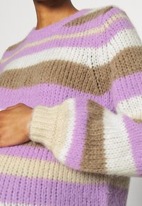 Osoba ubrana w dziergany sweter w poziome paski w kolorach lawendowym, beżowym i białym, ukazująca górną część tułowia i ramię.