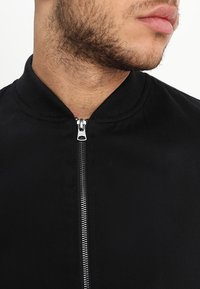 Černá bunda s uzavíráním na zip, žebrovaným límcem a hladkou strukturou. Obsahuje metalický zip a minimalistický design.