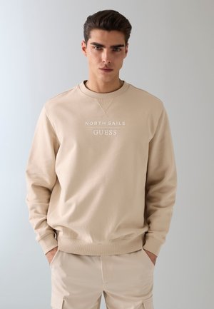 Młody mężczyzna o ciemnych włosach nosi beżową bluzę North Sails Guess i pasujące spodnie, stoi z rękami w kieszeniach na szarym tle.