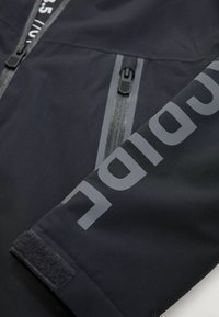 Manche de veste noire avec une fermeture éclair réfléchissante et le texte partiel "SPIDIR" en grandes lettres grises, plus une sangle en Velcro au niveau du poignet.
