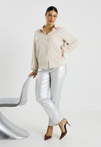 Blouse beige boutonnée avec manches longues, associée à un pantalon métallique argenté. Escarpins bordeaux pointus et coiffure lisse complètent le look.