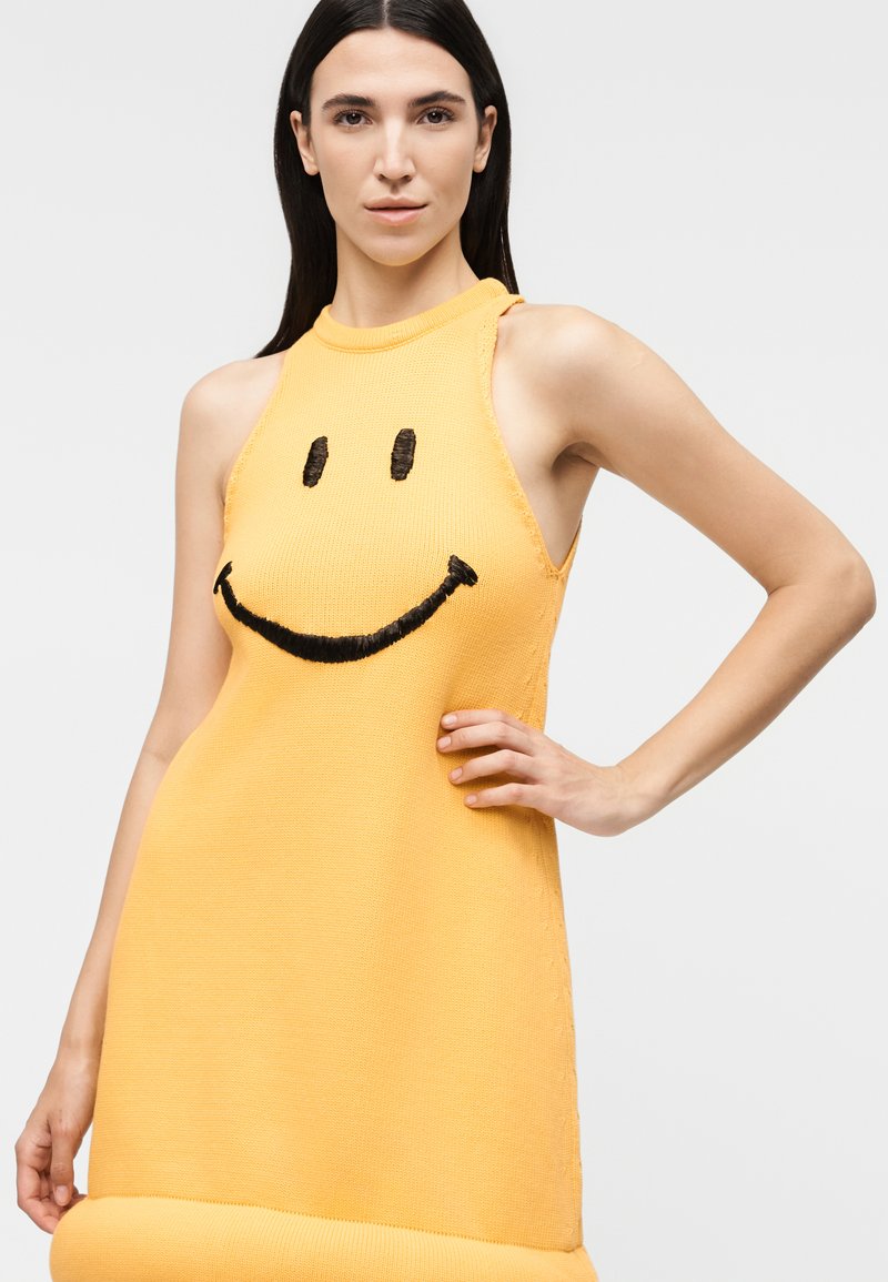 Woman with long dark hair wearing a sleeveless yellow dress featuring a large black smiley face, posing with one hand on her hip.