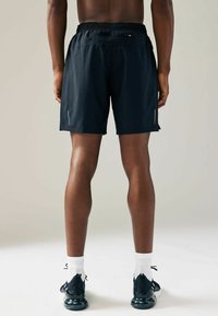 Shorts sportive negre cu talie elastică, buzunare laterale și accente verticale albe, prezentate din spate pe un fundal neutru.