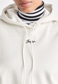 Hanorac crem cu șnururi, cu un logo brodat. Sub acesta, se observă un pulover cu guler înalt în dungi alb-negru. Textură de țesătură moale.