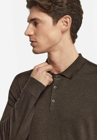 Brązowa, długorękawowa koszulka polo z kołnierzykiem, z trzema guzikami i ściągaczami na mankietach, pokazana na osobie z boku.