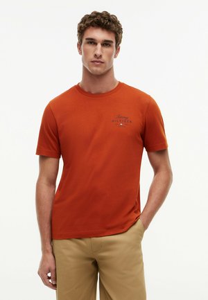 Joven con una camiseta Tommy Hilfiger de color naranja óxido y pantalones beige, de pie con una mano en el bolsillo contra un fondo blanco.