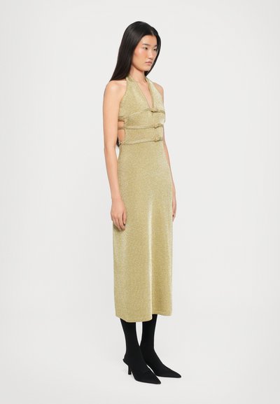 Sparkling gold halter dress with cut-out sides, featuring two button accents. Paired with black tights and pointed black heels.