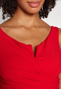 Abito rosso con scollatura a V e tessuto drappeggiato con una sottile texture. Il corpino è liscio, con linee morbide e fluide.