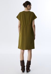 Frau, die mit dem Rücken zur Kamera steht, trägt ein olivgrünes knielanges Kleid mit kurzen Ärmeln und braune Stiefel bis zur Wade mit goldenen Schnallen.