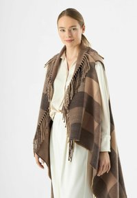 Poncho a scacchi marrone e beige con bordi frangiati, realizzato in tessuto morbido, indossato sopra una blusa color crema e pantaloni chiari.