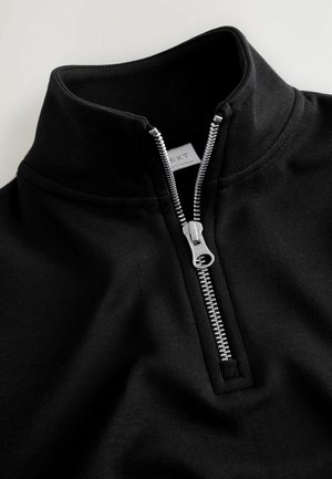 Sort sweatshirt med lynlås og høj krave lavet af struktureret stof. Har en sølv lynlås og et mærke synligt inden i halsudskæringen.
