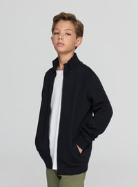 Czarna bluza na zamek z wysokim kołnierzem, wykonana z miękkiego materiału. Posiada długie rękawy i boczne kieszenie, noszona na białej koszulce.