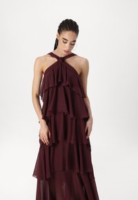 Bordo slojevita haljina napravljena od laganog materijala, s halter vratom i čvorom te naboranim slojevima, stvarajući laganu siluetu.