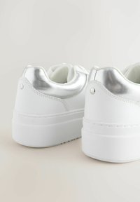 Scarpe da ginnastica bianche con accenti metallici argento, texture liscia, suole bianche elevate e dettagli di cucitura lungo la parte posteriore.