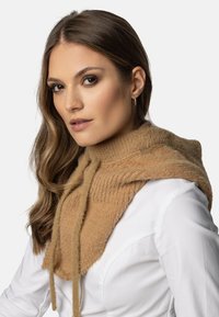 Să scarfă tricotată de culoare bej cu guler înalt, având o textură cordonată, șnururi și un material moale; purtată peste o bluză albă.