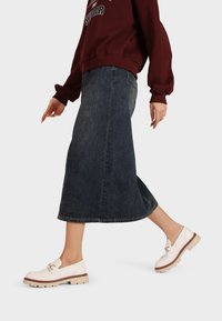 Scarpe slip-on bianche con suola beige e dettagli della cinghia, abbinate a una gonna midi in denim scuro e a una felpa bordeaux, evidenziando uno stile casual.