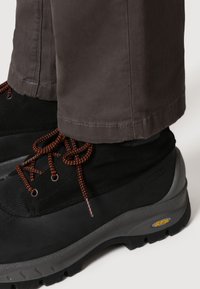 Svarta vandringsskor med orange snören, med en strukturerad överdel och grå sula med Vibram-märket. Byxor i en mörk nyans synliga ovanför.