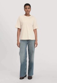 Cremfarbendes T-Shirt mit kurzen Ärmeln und Rundhalsausschnitt, kombiniert mit hellblauen, weit geschnittenen Jeans und braunen Schuhen, vor einem einfarbigen Hintergrund stehend.